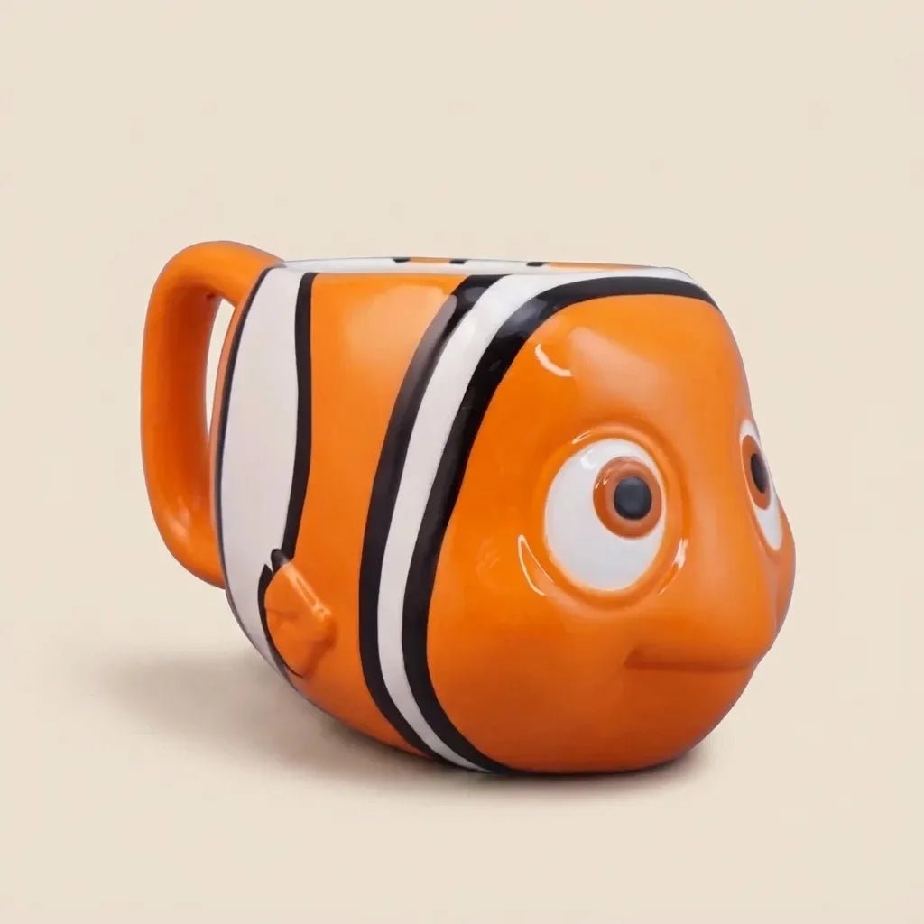 An orange clownfish-shaped ceramic mug with white and black stripes, designed to look like the character Nemo, on a solid beige background.