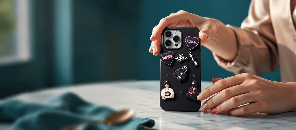 A person holds a black smartphone case decorated with multiple colorful patches and a pop socket, standing on a light marble tabletop with a blurred teal background.