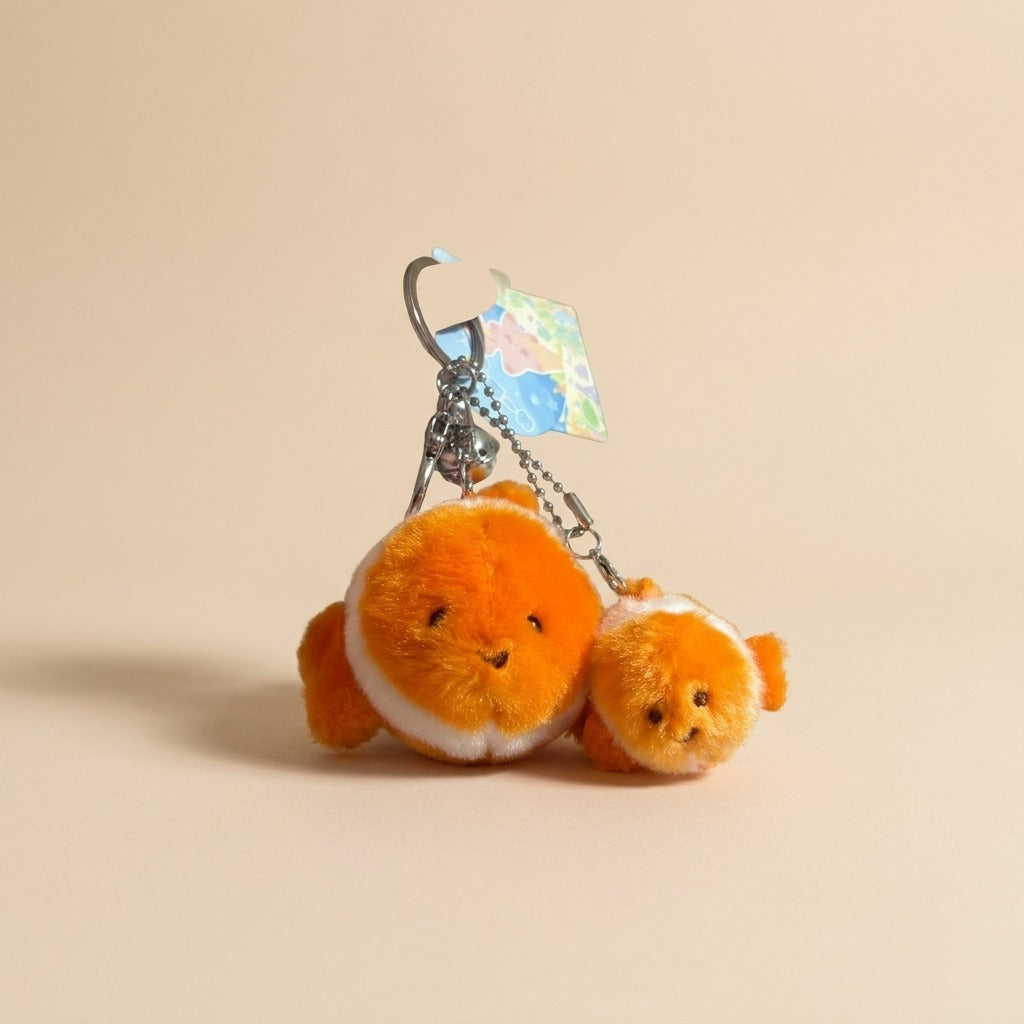 A plush keychain featuring two orange clownfish characters with white stripes, attached to a silver metal key ring and chain with a small map tag, set against a solid beige background.