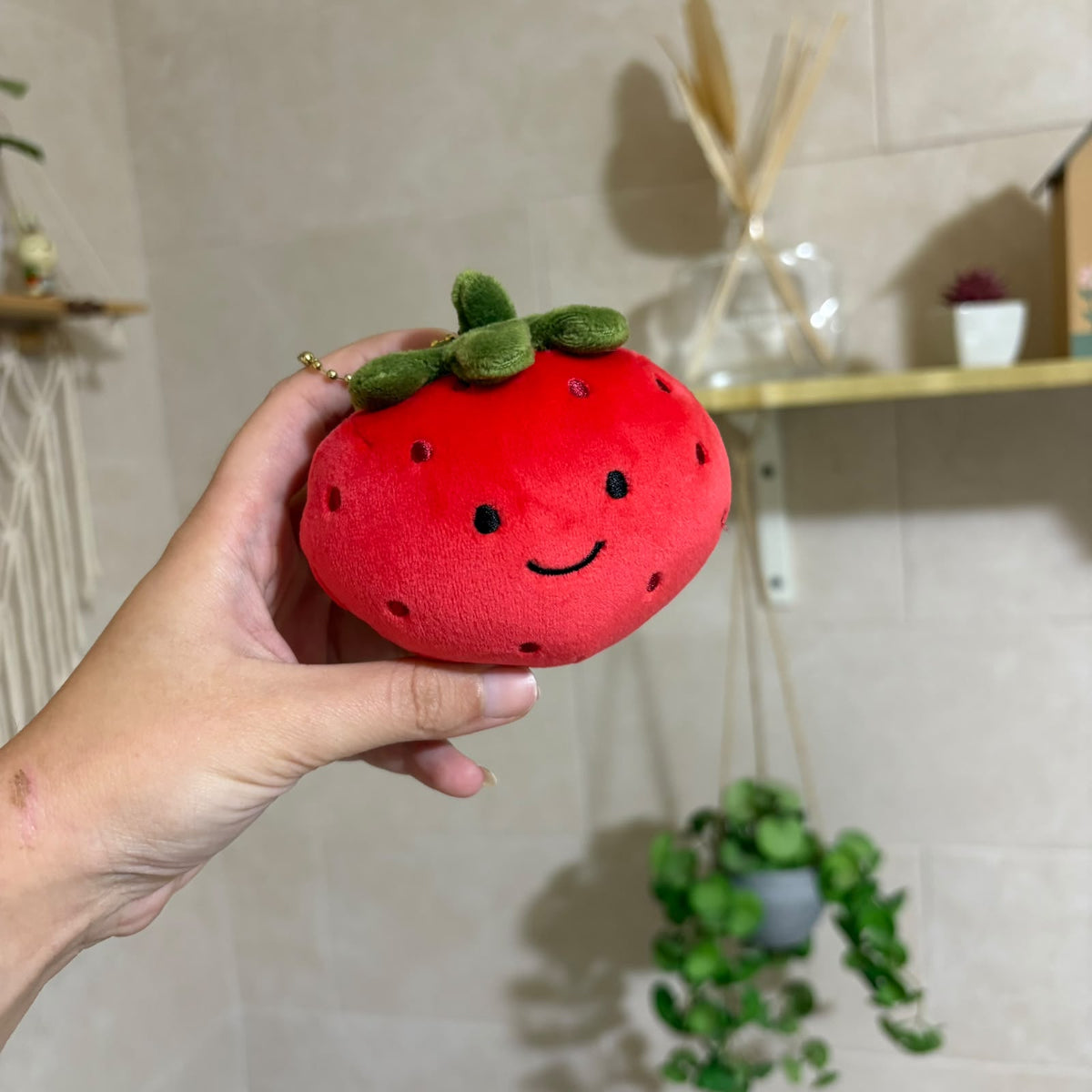 A hand holding a red strawberry plush toy with green leaves and a smiling face, set against a softly blurred indoor background with shelves and plants.