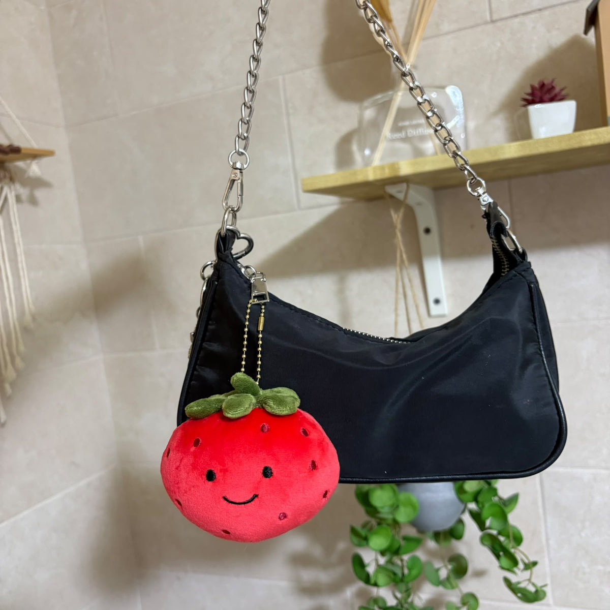 A black nylon shoulder bag with a silver chain strap, featuring a cute red strawberry plush keychain charm with green leaves and a smiling face, hanging against a beige tiled wall.