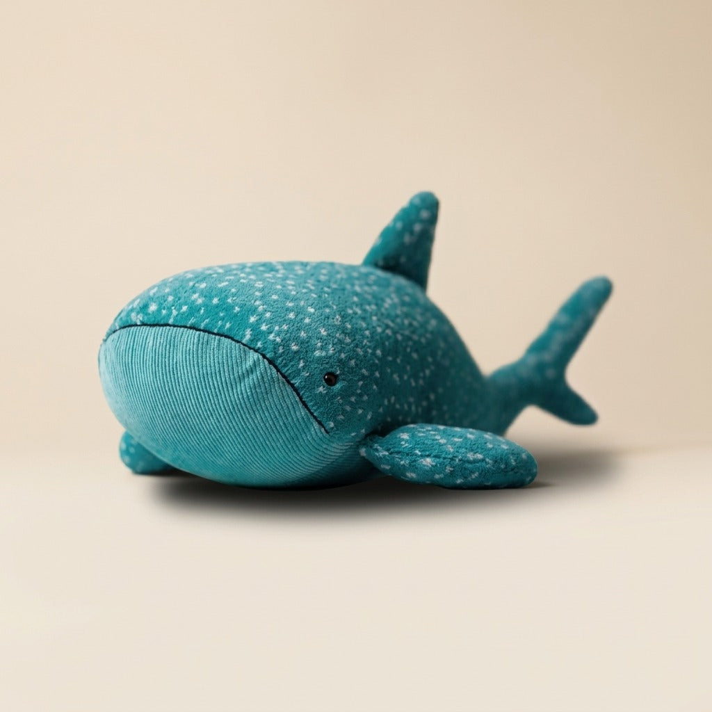 A teal blue whale-shaped plush toy with white speckles, photographed on a clean, solid beige background.
