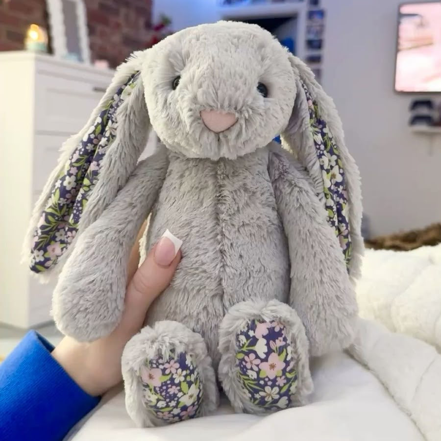 A gray plush bunny stuffed animal with floral-patterned ears and feet, held by a hand on a white bed in a bedroom setting.
