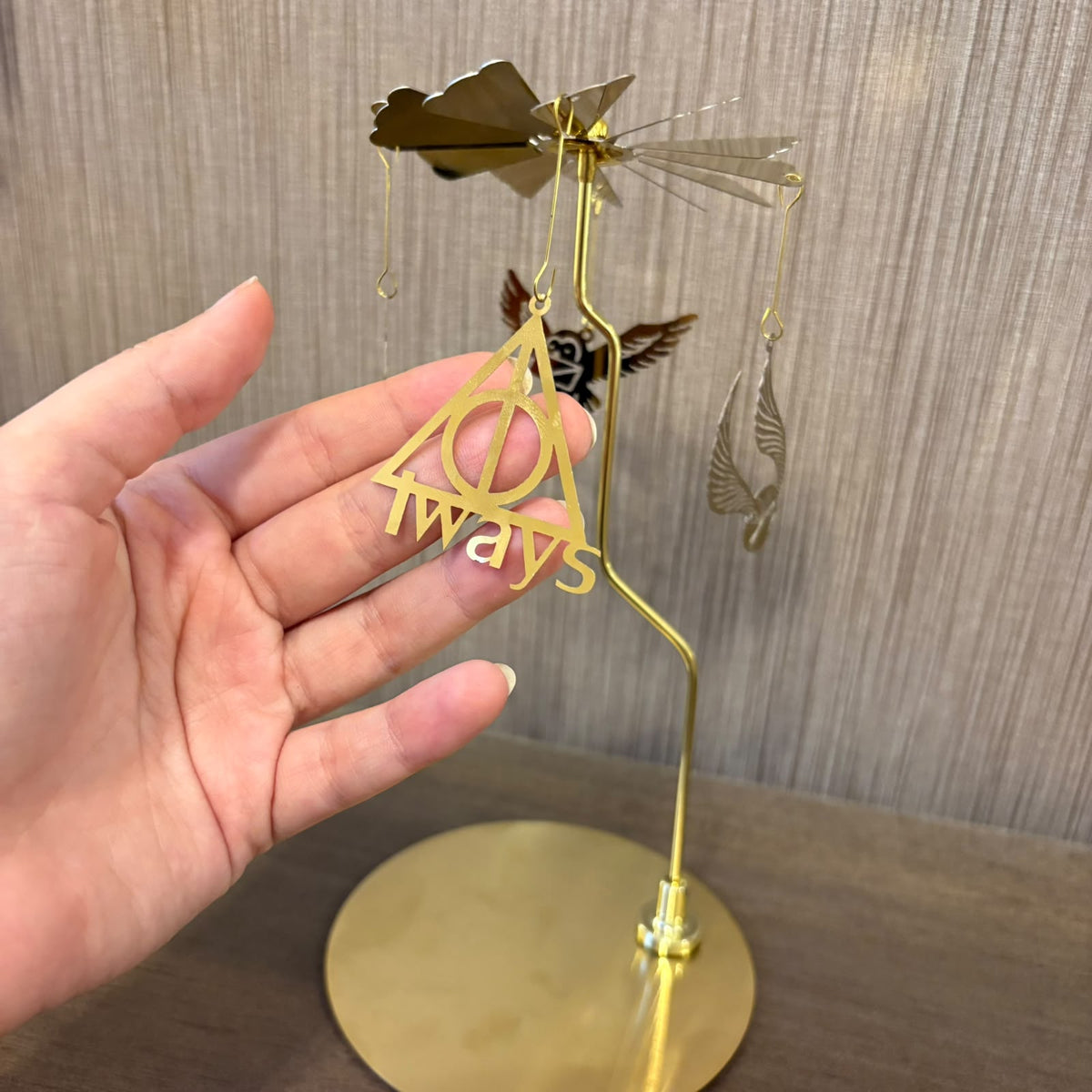 A hand holds a gold-colored metal wind spinner ornament featuring the Death Star symbol and the word 'Always' from Star Wars, with a dragonfly and feather details, set against a wooden background.