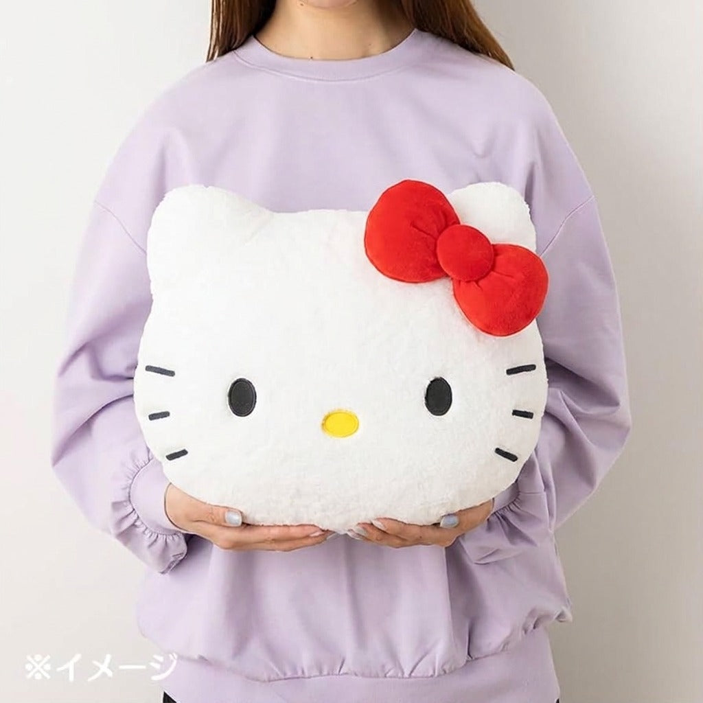A person wearing a light purple sweatshirt holds a white Hello Kitty face plush pillow with a red bow and yellow nose against a plain light background.