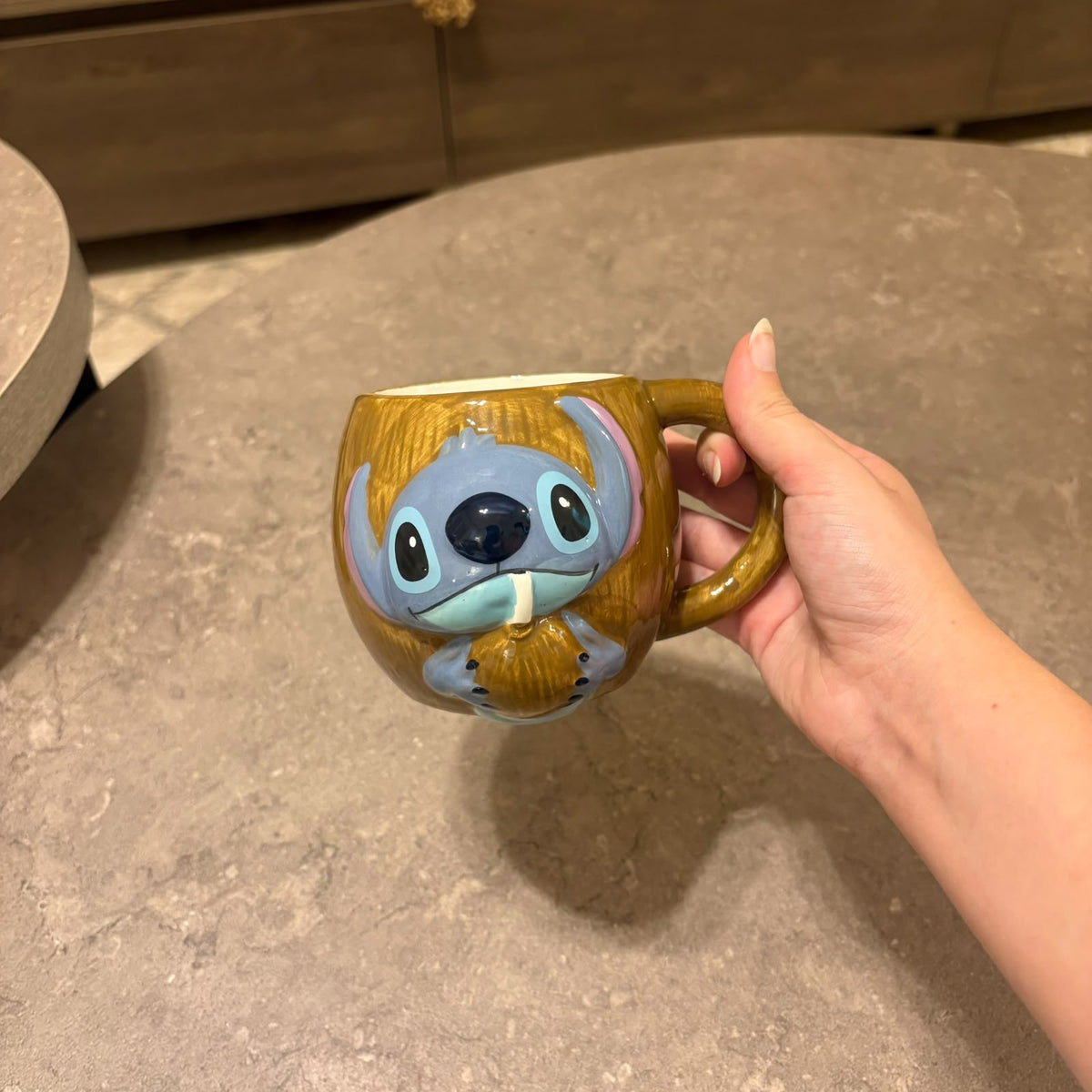 A hand holding a brown ceramic mug featuring a 3D cartoon character with blue-gray skin and large black eyes, set against a gray stone tabletop.