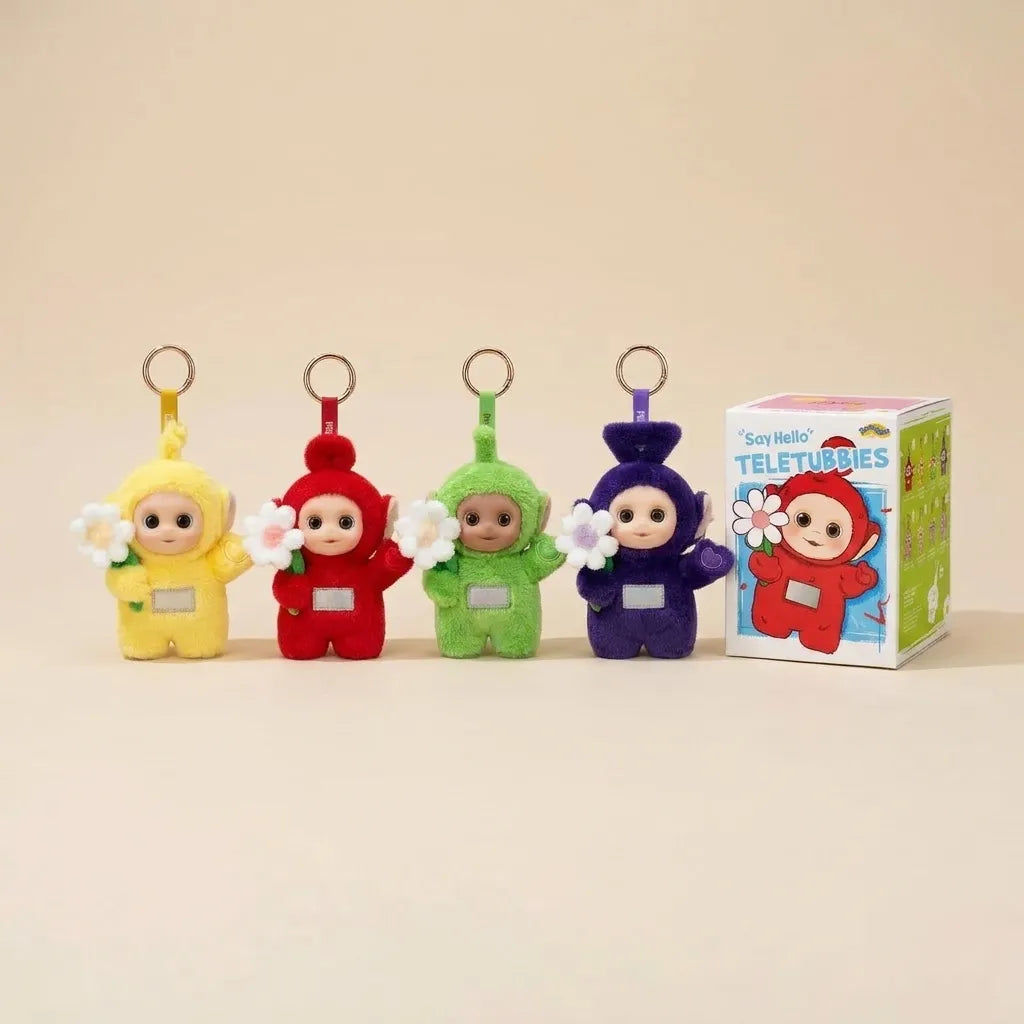 Four Teletubbies plush keychains in yellow, red, green, and purple, each holding a white flower, displayed next to their branded retail box on a warm beige background.