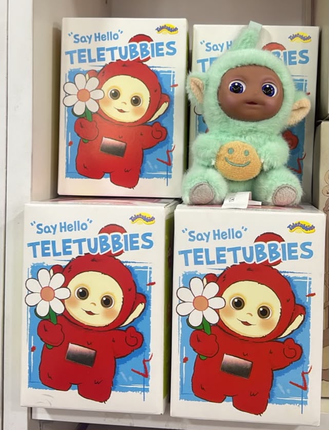 Three 'Say Hello Teletubbies' boxes arranged on a shelf, with a plush Teletubby toy in a mint green outfit placed on top of the right box.