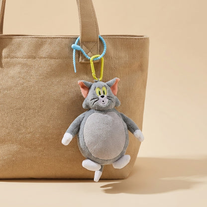 A plush character keychain shaped like a gray cat with pink ears, yellow eyes, and white paws, hanging from a blue and yellow clip on a beige canvas tote bag against a warm beige background.