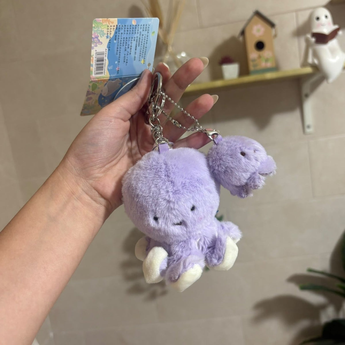 A hand holds a lavender plush octopus keychain with a silver key ring and a matching small plush piece attached. The keychain has a paper tag still attached, and the background shows a beige tiled wall with a shelf holding small decorative items.