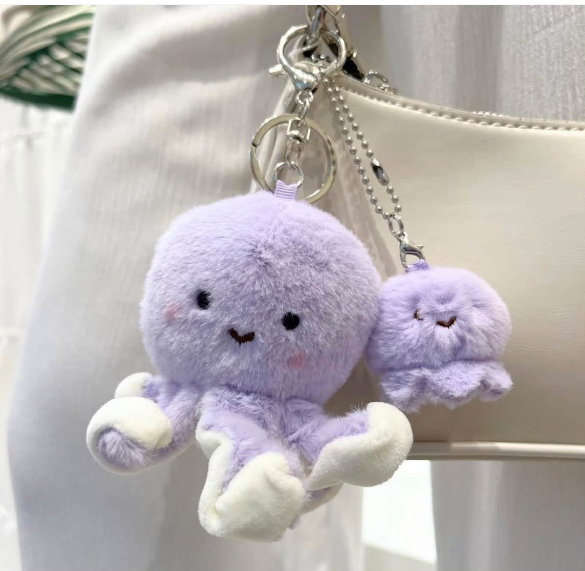 A cute lavender plush octopus keychain with a matching small charm, hanging from a silver key ring and chain next to a cream-colored handbag.