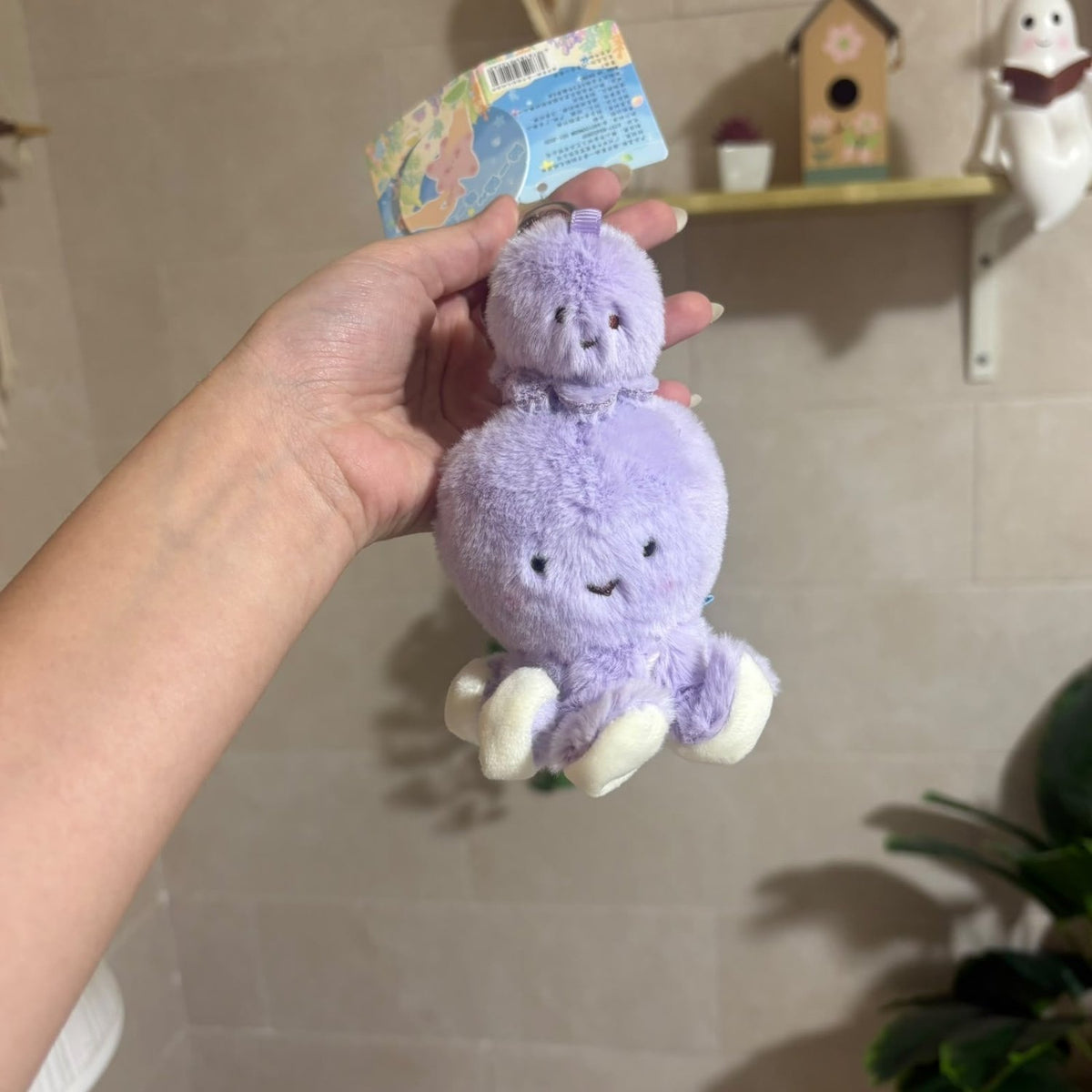 A hand holds a small purple plush octopus keychain with white feet and a colorful hang tag, photographed indoors against a beige tiled wall with shelf decor in the background.