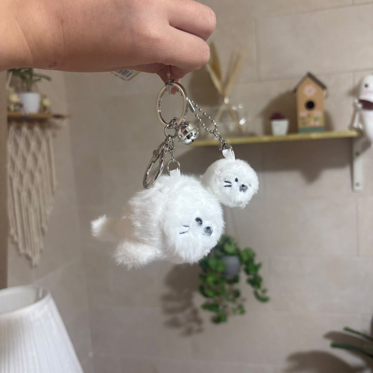 A hand holds a keychain with two fluffy white cat-shaped charms and small silver bells in a home setting with a beige tiled wall and shelf in the background.