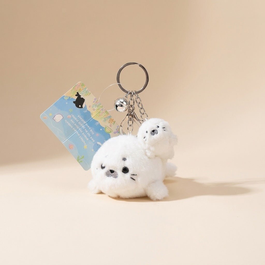 A white plush seal keychain with a small attached card and metal key ring, photographed on a warm beige studio background.