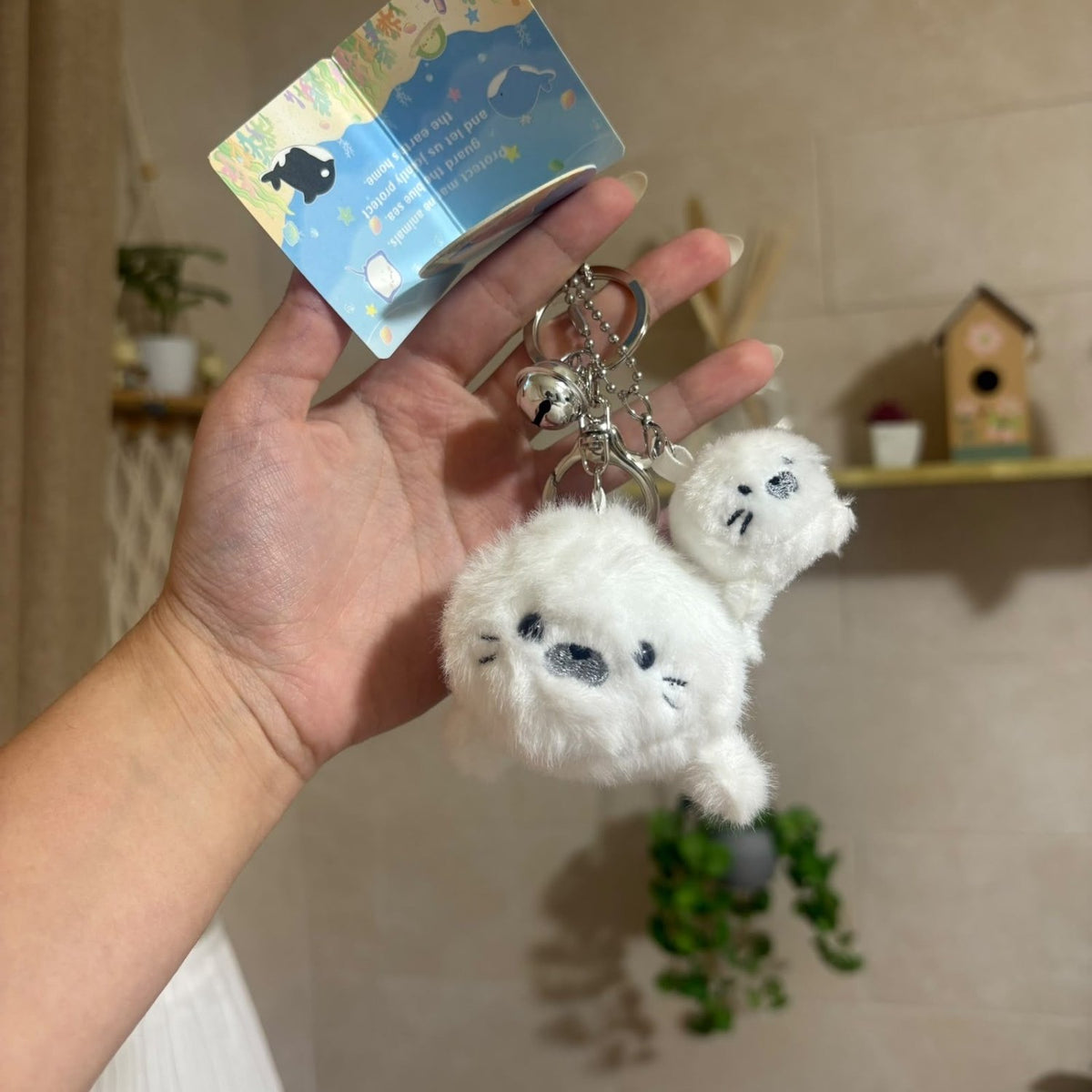 A hand holds a white plush seal keychain with a small attached plush and a colorful paper tag in a home setting with a tiled wall and shelf in the background.