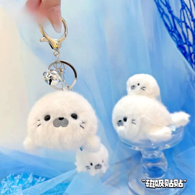 A white plush seal keychain charm with a gold clasp and silver bell, shown with smaller matching plush seal stickers on a blue tulle backdrop.