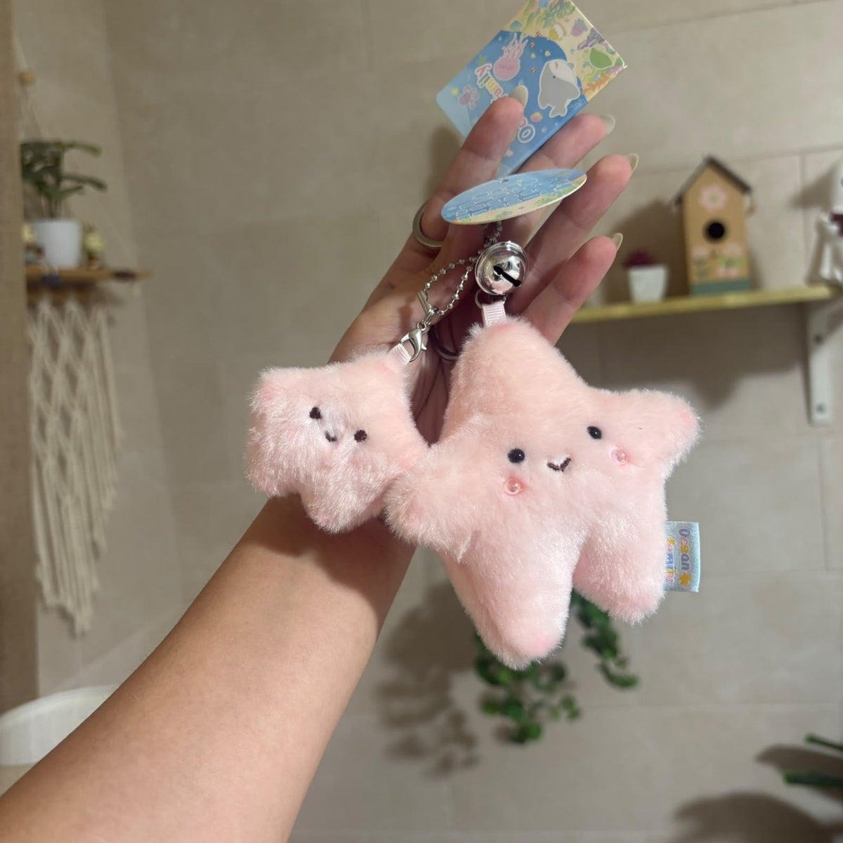 A person's hand holds a pink plush star-shaped keychain charm with a small silver bell and tags, set against a softly blurred indoor background with shelves and plants.