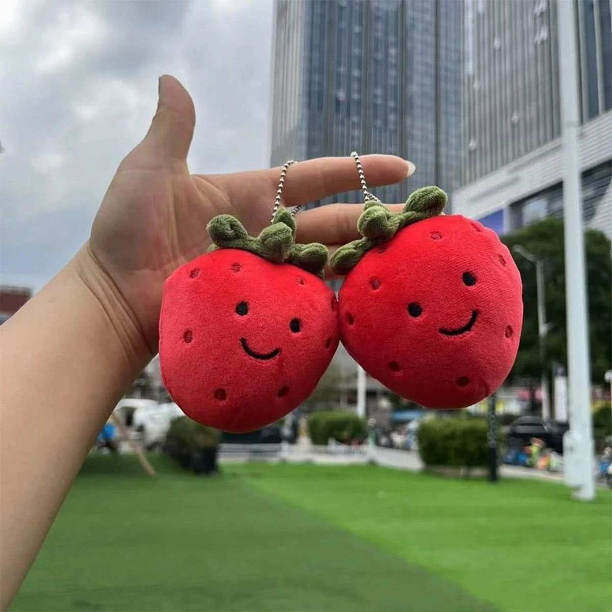 Two red strawberry plush keychains with embroidered smiling faces and green leaf tops are held in a person's hand outdoors, with a blurred city park and buildings in the background.