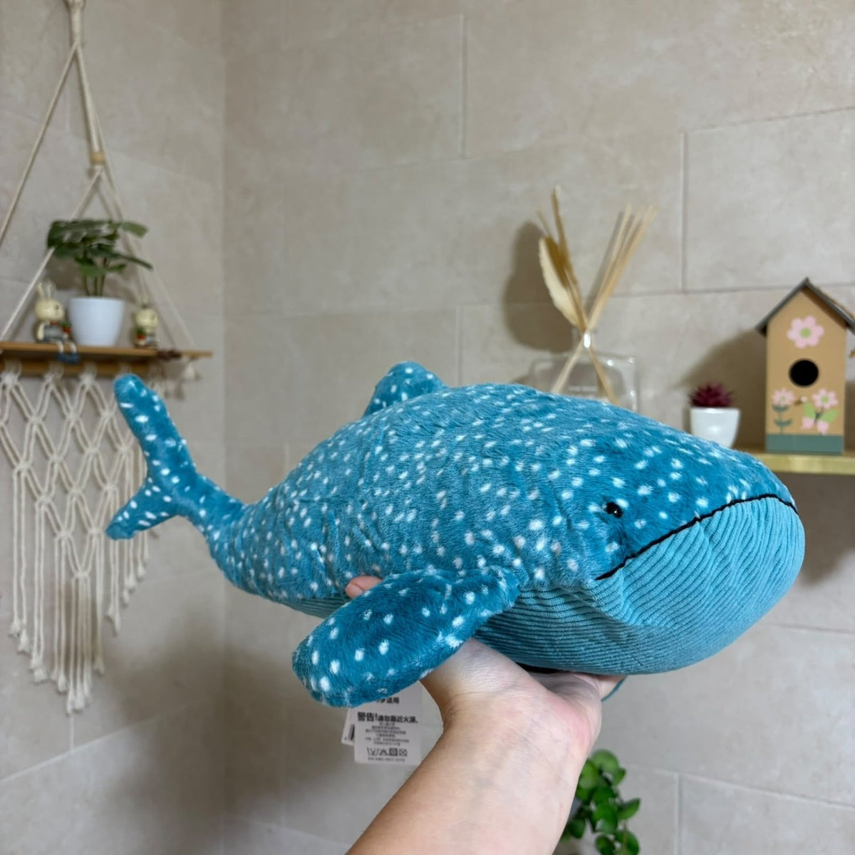 A plush whale shark toy being held up by a person's hand in a home setting with beige tiled walls, a macrame wall hanging, and small decor items in the background.