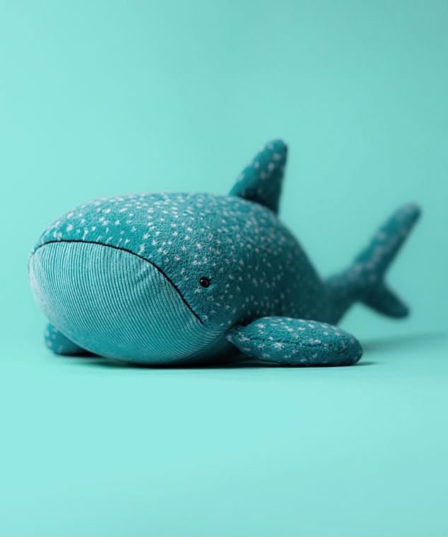 A plush whale shark toy made of textured teal-blue fabric with white polka dots, resting on a solid teal background.