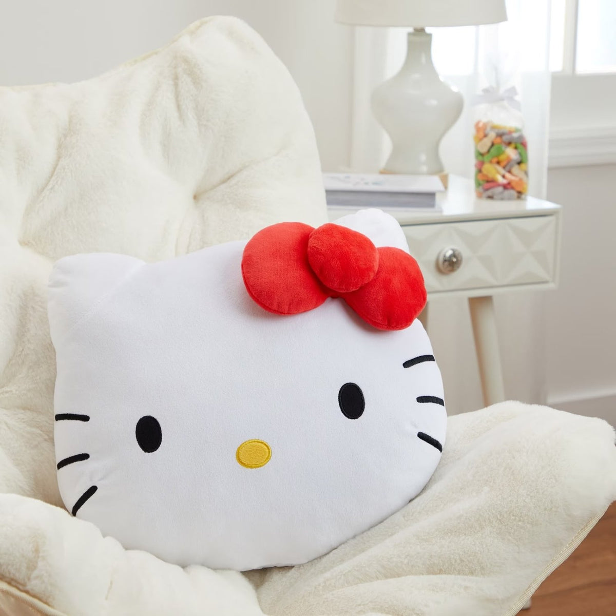A Hello Kitty plush pillow with a white face, black whiskers and eyes, and a red bow, resting on a cream-colored armchair in a bright room.
