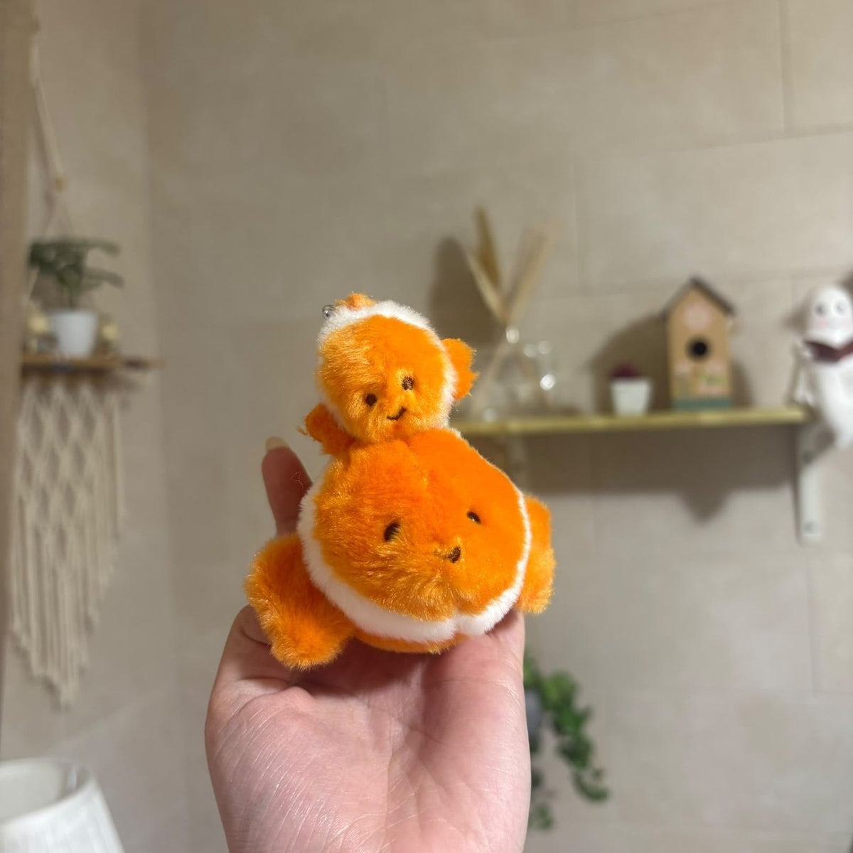 A small orange and white plush fish keychain held in a person's hand, with a softly blurred indoor background showing shelves and decor.