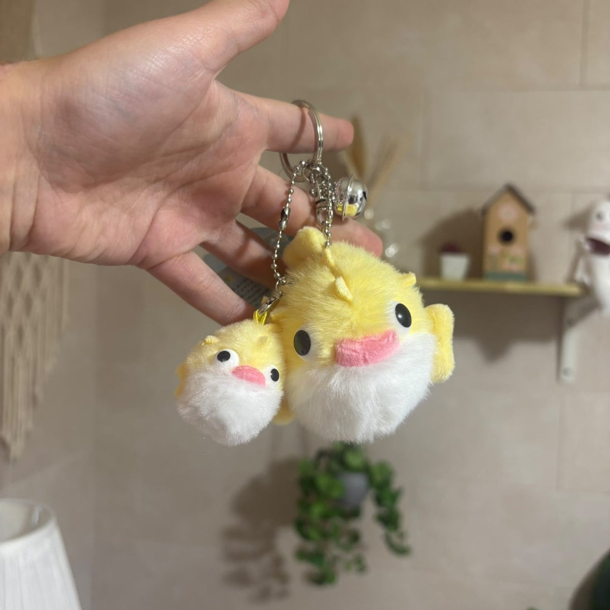 A hand holds two small plush pufferfish keychains, one yellow and one white, with silver keychains and a small bell attached, against a softly blurred indoor background.