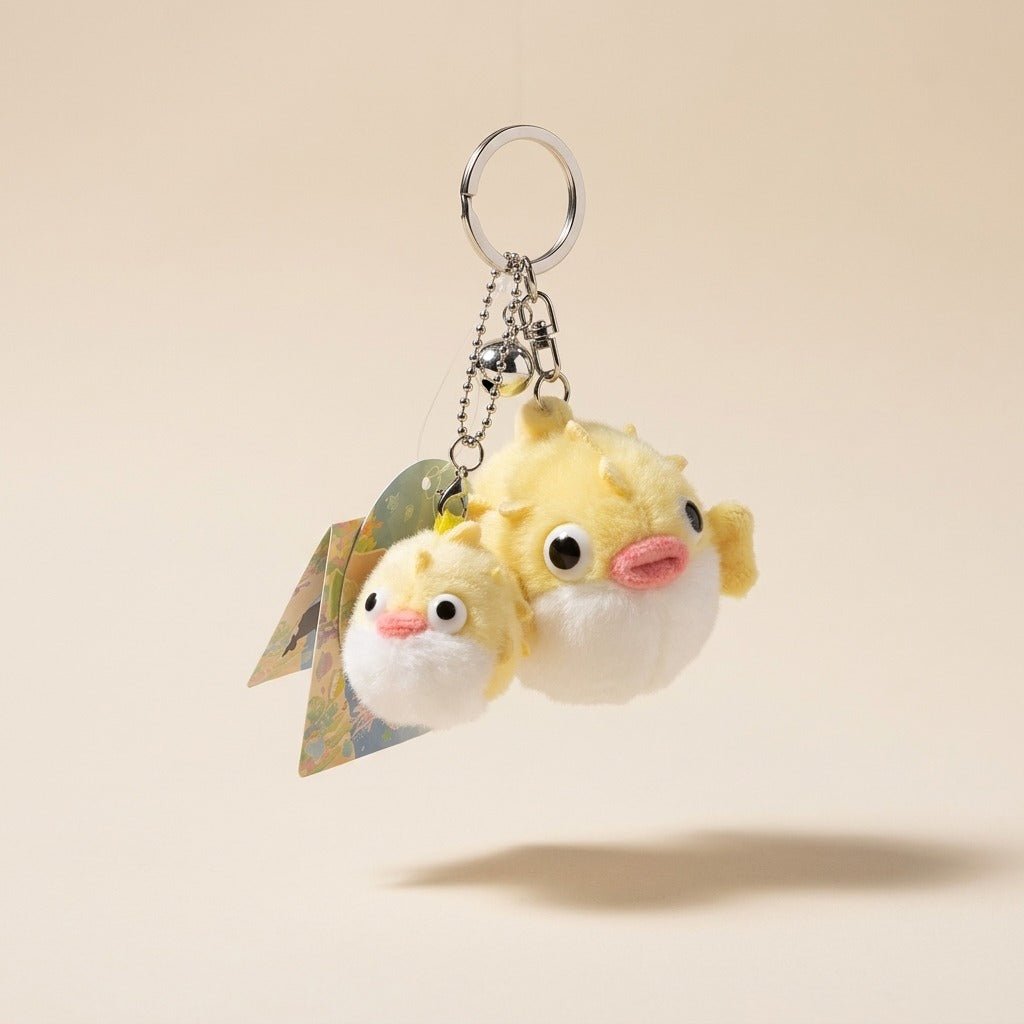 A plush puffer fish keychain featuring two small yellow and white fish characters with pink noses, attached to a silver metal key ring and ball chain, shown against a warm beige background.