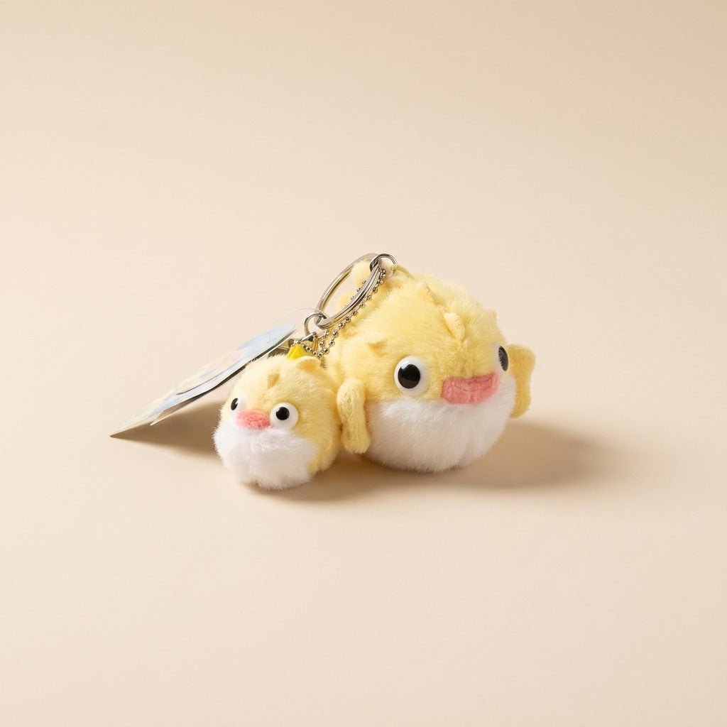 A yellow plush keychain shaped like a cute bird character with a silver metal key ring and a small paper tag, photographed on a plain warm beige background.