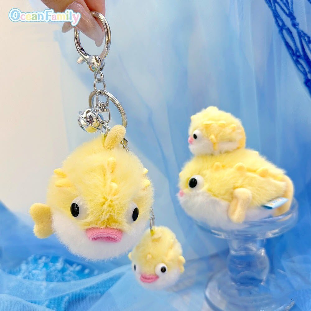 A yellow plush duck keychain with a silver metal key ring and bell, held by a hand. The keychain features a cute duck face with black eyes and a pink beak.