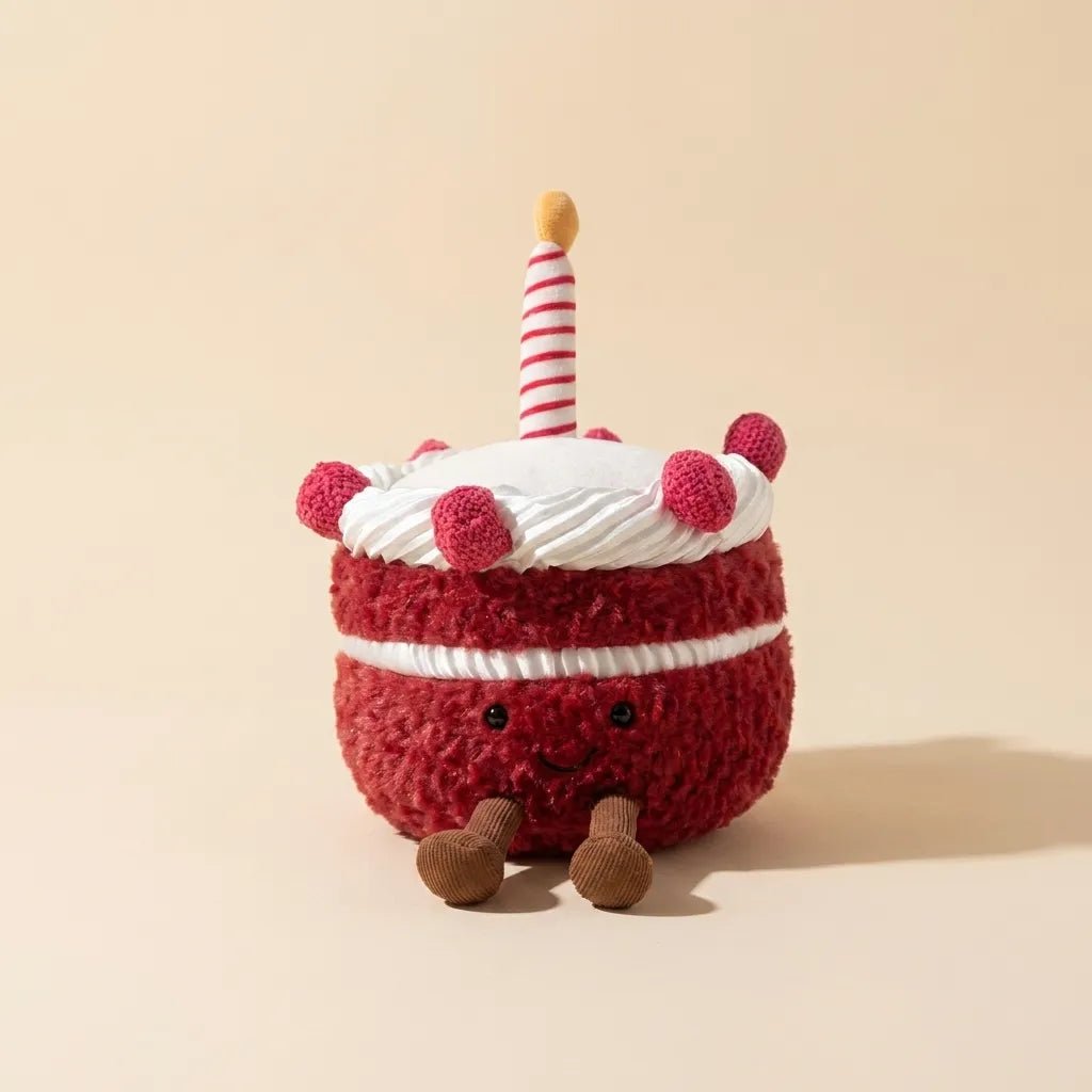 A plush cake toy with red fuzzy frosting, white frosting, a striped candle, and small brown legs, photographed on a warm beige studio background.