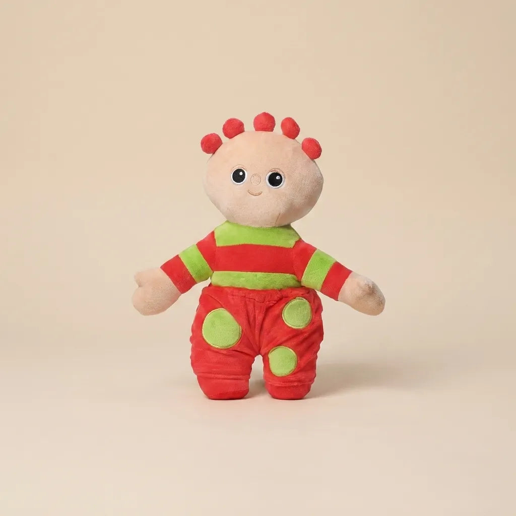 A small plush toy character standing against a plain warm beige studio background. The character has a cream face and hands, red hair spikes, a red-and-green striped top, and red pants with green circular patches.