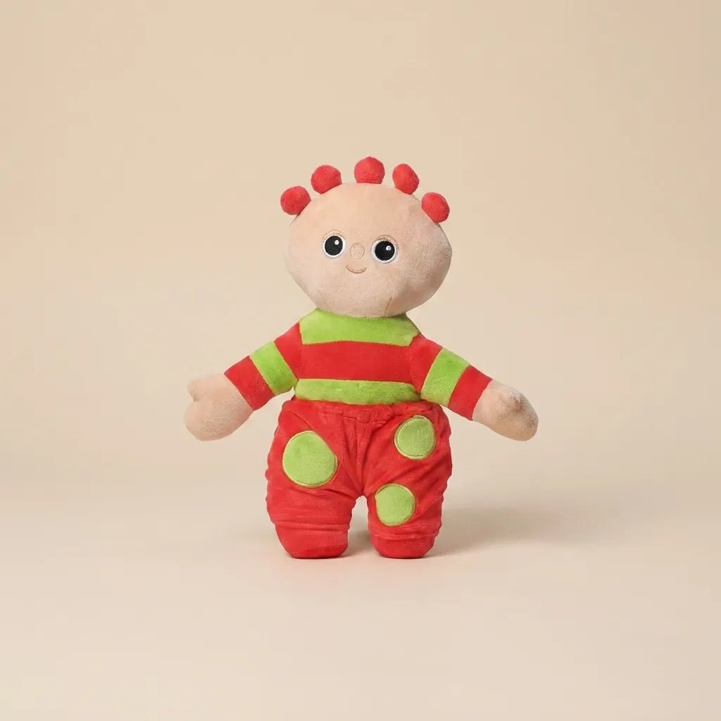 A small plush toy character standing against a plain warm beige studio background. The character has a cream face and hands, red hair spikes, a red-and-green striped top, and red pants with green circular patches.